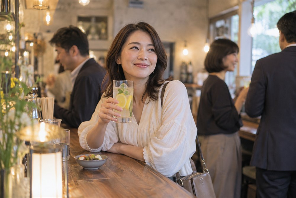 明るい立ち飲み屋でソフトドリンクと料理を楽しむ一人客の女性のイメージ